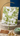 Decorative pillow with green floral patterns on a wooden stand, with earrings and a card in the foreground.